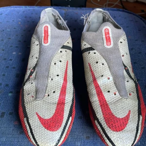 Nike phantom gt fotbollsskor  med röd och silver design Elite - Säljer ett par Nike Mercurial Vapor fotbollsskor i silver, rött och svart med stora röda swoosh-loggor. Skorna har en strömlinjeformad design och plös i grått. Perfekta för snabba spelare som vill sticka ut på planen.