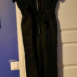 Svart lång jumpsuit med dragkedja och knytband - Säljer en svart lång jumpsuit med korta ärmar, dragkedja framtill och knytband i midjan. Perfekt för dig som vill ha en stilren och bekväm look. Passar lika bra till sneakers som till klackar. Aldrig använd
