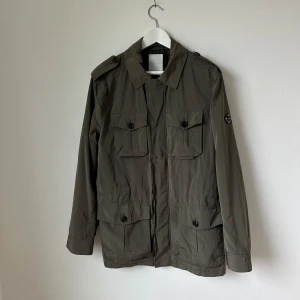 Olivgrön Foreman jacka från J.Lindeberg - Säljer en olivgrön jacka från J.Lindeberg med fyra stora fickor framtill och axelklaffar. Jackan har lång ärm, knappar och dragkedja samt klassisk krage. Perfekt för lager-på-lager under kyliga dagar. Modell: Foreman 62 Peach Memo - Artnr: Ä248