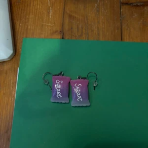 Rosa och lila örhängen med texten 'Sweet' - Säljer ett par unika örhängen i form av små påsar med texten 'Sweet' i vitt. Örhängena har en snygg färgövergång från rosa till lila och är perfekta för dig som vill sticka ut med din stil.