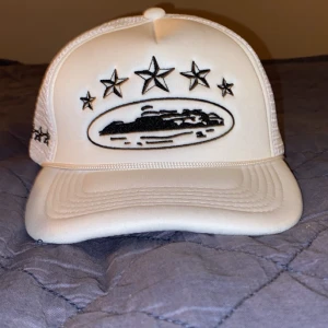Vit truckerkeps med broderade stjärnor - Snygg vit truckerkeps från Corteiz med svart broderad grafik framtill, fem stjärnor och ett motiv i mitten. Sidorna är i mesh för extra ventilation och bak finns justerbart spänne. Broderad logga baktill.