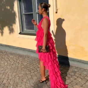 Rosa volangklänning med djup v-ringning (NA-KD) - Säljer min rosa klänning med flera lager volanger och djup v-ringning, klänningen är endast använd av min vän under fotografering. Klänningen har ärmlös design och asymmetrisk längd med lång volang baktill. Är i perfekt skick. Storlek XXS. Passar en XS-S då den är stor i storlek och lång. 