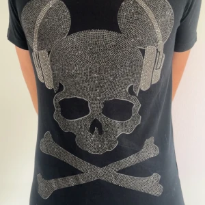 Svart t-shirt med döskalle och hörlurar - Cool svart t-shirt med glittrigt döskalletryck med hörlurar på framsidan och vit detalj i ryggen. Perfekt för dig som gillar unika och edgy plagg. Kortärmad och normal passform. Storleken är S men passar M