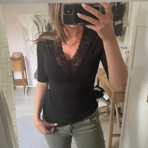 Svart topp med spets från By Malene Birger - Säljer en svart topp från By Malene Birger med v-ringning och fina spetsdetaljer vid halsen. Toppen har korta ärmar och en figurnära passform. Perfekt att styla till olika outfits.