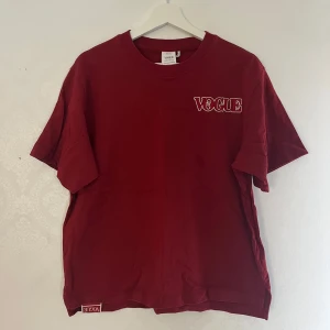 Röd t-shirt från Puma x Vogue - Snygg röd t-shirt från Puma i samarbete med Vogue. T-shirten har korta ärmar, normal passform och tryckt Vogue-logga på bröstet. Har en fläck på framsidan som man kan se på bild 3.  