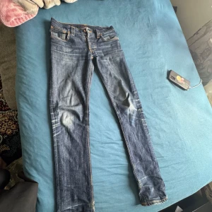 Mörkblå jeansbyxor från Nudie Jeans - Säljer ett par mörkblå jeans från Nudie Jeans med klassisk femficksdesign och orangea sömmar. Byxorna har en rak passform och normal midja, perfekta till vardags. Nudie-logga på bakfickan och läderpatch bak i midjan.