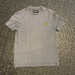 Grå t-shirt från Lyle & Scott - Säljer en stilren grå t-shirt från Lyle & Scott med klassisk gul logga på bröstet. T-shirten har rund halsringning och korta ärmar. Perfekt basic-plagg till garderoben.