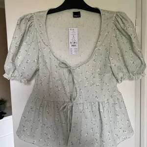 Ljusgrön blus med knyt  - Supersöt ljusgrön blus från Gina Tricot med små blommiga detaljer och knyt framtill. Blusen har puffiga korta ärmar och en lätt volang nedtill. Perfekt för vår och sommar!