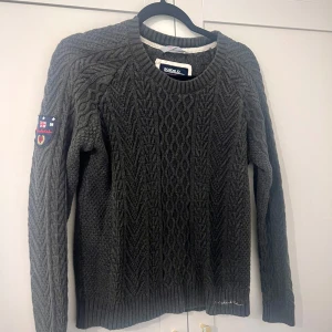 Mörkgrå stickad tröja från Bondelid - Säljer en mörkgrå stickad tröja från Bondelid med klassiskt kabelstickat mönster. Tröjan har rund halsringning, långa ärmar och en patch på ena ärmen. 