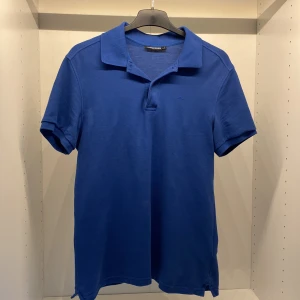 Blå piké t-shirt från J.Lindeberg - Snygg blå piké t-shirt från J.Lindeberg med klassisk krage och knappslå. T-shirten är kortärmad och har en diskret broderad logga på bröstet. 