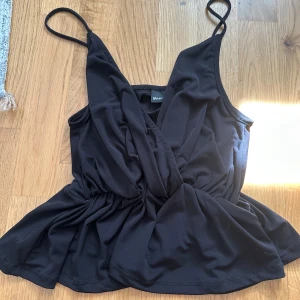 Svart omlottopp från Gina Tricot - Säljer en svart omlottopp från Gina Tricot i storlek XS. Toppen har smala axelband, omlottdesign och markerad midja som ger en snygg siluett. Perfekt till sommaren och enkel att matcha med olika underdelar.