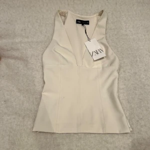 Vit ärmlös topp från Zara - Säljer en stilren vit ärmlös topp från Zara med djup v-ringning och figurnära passform. Helt oanvänd! så så fin och passar till allt. Säljer då jag tyvärr inte fått användning av den. Prislapp är kvar så skicket är som nytt.🤗