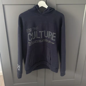For the culture hoodie  - Säljer en mörkblå hoodie med texten 'FOR THE CULTURE'. Tröjan har huva, magficka och långa ärmar. Perfekt för en avslappnad stil och enkel att matcha med jeans eller joggers. Aldrig använd