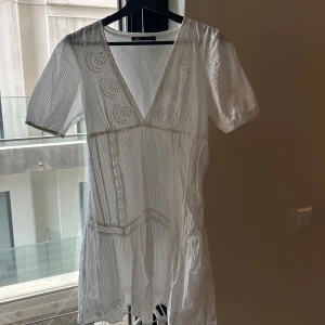 Vit kortklänning med spetsdetaljer - Säljer en söt vit kortklänning med v-ringning och fina spetsdetaljer. Klänningen har korta ärmar och lätt utställd kjol. Perfekt för varma dagar eller speciella tillfällen.