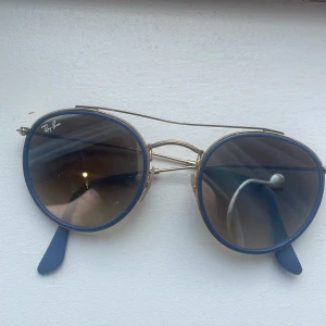 Runda solglasögon från Ray-Ban med guldfärgade bågar - Snygga runda solglasögon från Ray-Ban med tunna guldfärgade metallbågar och mörktonade glas. Skalmarna har blå detaljer längst ut. Perfekt accessoar för soliga dagar.