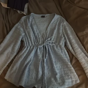 Ljusblå omlottopp från Gina Tricot - Säljer en ljusblå omlottopp från Gina Tricot med lång ärm och knyt i midjan. Toppen har ett vågigt, strukturerat tyg och en v-ringad urringning. Säljer pga för stor storlek🤍