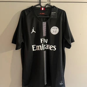 Svart Paris Saint-Germain fotbollströja - Säljer en svart PSG fotbollströja med vita detaljer och Fly Emirates-tryck på bröstet. Tröjan har korta ärmar, Paris Saint-Germain klubbmärke och Jordan-logga framtill. Tillverkad i lätt och ventilerande material, perfekt för träning eller match.