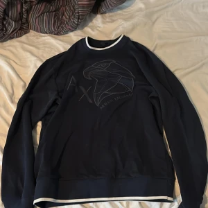 Mörkblå sweatshirt från Armani Exchange - Säljer en mörkblå sweatshirt från Armani Exchange med broderad örn och logga på bröstet. Tröjan har vita detaljer vid hals, ärmslut och nederkant. Perfekt för en stilren och sportig look.