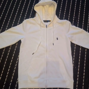 Vit hoodie från Polo Ralph Lauren - Säljer en klassisk vit hoodie från Polo Ralph Lauren med dragkedja och huva. Tröjan har det ikoniska broderade logotypen på bröstet och justerbara snören i huvan. Perfekt för en stilren och avslappnad look. Finns en liten matfläck på tröjan som borde försvinna eller minskas efter tvätt, priset kan diskuteras.