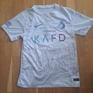 Al Nassr FC Ronaldo 7 fotbollströja - Säljer en vit Al Nassr FC fotbollströja från Nike med Ronaldo 7 på ryggen. Tröjan har korta ärmar, klubbmärke och sponsorloggor på framsidan samt färgade detaljer i blått och lila. Perfekt för fans av fotbollsfans!