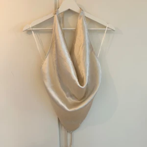 Champagnefärgad satintopp med drapering - Super fin topp med halterneck och öppen rygg.  Köpt från Gina tricot för några år sen men aldrig kommit till användning pågrund av fel storlek. Skickar gärna fler bilder om så önskas då bilderna inte visar toppen jätte bra! 