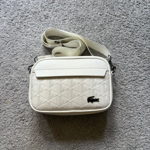 Vit axelväska från Lacoste - Snygg vit axelväska från Lacoste med quiltad framsida och det klassiska krokodilmärket i metall. Väskan har justerbar axelrem och stilren design med geometriskt mönster på framsidan. Perfekt för att bära dina essentials med stil.