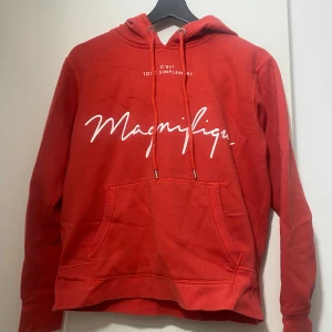 Röd hoodie med tryck från H&M - Röd hoodie från H&M med vit text 'Magnifique' framtill. Tröjan har huva med snörning, känguruficka och långa ärmar. Skön och avslappnad passform, perfekt för chill dagar. Tillverkad i mjuk bomullsmix.