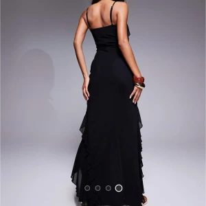 Svart långklänning med volangdetaljer - Supersnygg svart långklänning med tunna axelband och v-ringning. Klänningen har volangdetaljer längs sidorna och en slits framtill som ger en extra touch. Materialet är lätt och faller snyggt längs kroppen, perfekt för en elegant look. Aldrig använd, taggen på!!!