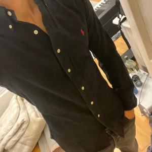 Svart skjorta från Ralph Lauren - Säljer en svart långärmad skjorta från Ralph Lauren med klassisk passform. Skjortan har vita knappar och den ikoniska röda logotypen broderad på bröstet. Tillverkad i mjuk bomull, perfekt för en clean och stilren look. Stilren skjorta för sommaren 