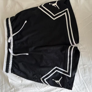 Svarta Jordan shorts med vita detaljer - Snygga svarta Jordan shorts med vita ränder och logga på benen. De har elastisk midja med snörning och är tillverkade i ett ventilerande meshmaterial. Perfekta för sport eller chill, med en loose fit och sportig vibe.