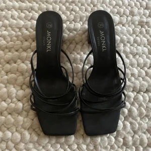 Svarta sandaler med klack från Monki - Stilrena svarta sandaler från Monki med fyrkantig tå och tunna remmar. Skorna har en låg klack och är tillverkade i veganskt syntetmaterial. Perfekta för dig som gillar minimalistisk och trendig design.