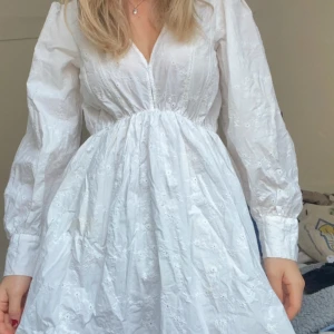 Vit broderad klänning med v-ringning - Superfin vit klänning med broderade blommor, v-ringning och långa puffärmar. Klänningen har markerad midja och är gjord i bomull, perfekt för sommarens alla tillfällen. Den har en romantisk och luftig känsla med fina detaljer.