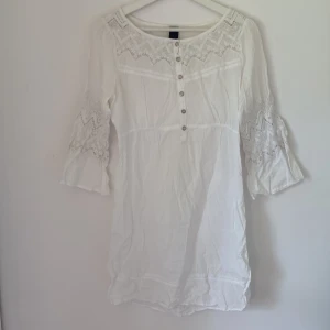 Vit spetsklänning från Vila, S - Superfin vit kortklänning från Vila med spetsdetaljer på ärmar och över bröstet. Klänningen har trekvartsärm, knappar framtill och en lätt boho-vibe. Perfekt för sommarens alla tillfällen och riktigt skön passform.