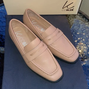 Beiga loafers från K.Cobler, stl 38 - Snygga loafers i beige skinn från K.Cobler. Klassisk design med markerad söm och låg klack. Skorna har en stilren pennyloafers-detalj och svart sula. Perfekta för dig som gillar en clean och tidlös look. De är helt nya endast testade hemma. Nypris 999kr. 