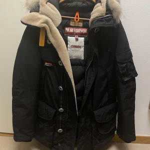 Svart dunjacka Parajumpers med päls - Säljer en svart Parajumpers dunjacka med stor huva och fluffig pälsdetalj. Jackan har flera fickor, coola badges på ärmen och orange detaljer. Perfekt för kalla dagar med sin tjocka vaddering och snygga design. Dragkedja och knappar framtill. Använd fåtal gånger som ny. nypris 11999