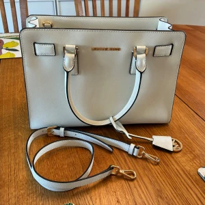 Vit handväska från Michael Kors - Snygg och stilren vit handväska från Michael Kors i skinn med guldiga detaljer. Väskan har både handtag och avtagbar axelrem, samt ett rymligt fack med dragkedja inuti. Klassisk fyrkantig form och diskret logga framtill. Äkta och oanvänd (avskedspresent från kollegor).