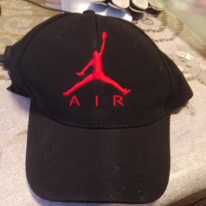 Svart Air Jordan keps med röd logga - Svart keps från Air Jordan med den klassiska röda Jumpman-loggan och texten AIR broderad framtill. Justerbar rem baktill med metallspänne. Tillverkad i 100% polyester, perfekt för en sportig och streetinspirerad stil.