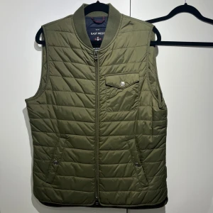 Olivgrön quiltad dunväst East West - Quiltad olivgrön dunväst från East West med dragkedja framtill, ribbad krage och tre fickor med knappdetaljer. Perfekt för lager-på-lager och snygg till både hoodie och t-shirt.