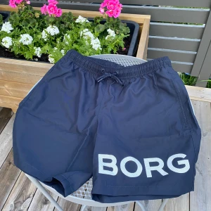 Badshorts från Björn Borg - Svarta badshorts från Björn Borg med stor vit logga på ena benet. Shortsen har elastisk midja med snörning och praktiska fickor. Perfekta för stranden eller poolen, tillverkade i snabbtorkande syntetmaterial. Alldrig annvända då de är för små. Kvitto finns från XXL. Priset är ej hugget i sten. Skriv vid minsta lilla fundering  