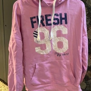 Rosa hoodie från Atmosphere - Rosa hoodie med tryck 'FRESH 96' i vitt och mörkblått framtill. Tröjan har huva med vita snören och en stor magficka. Mjuk och skön, perfekt för chill dagar. Långärmad och i klassisk passform.