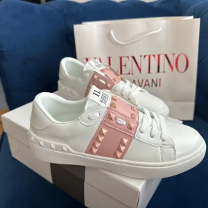 Valentino Rockstud sneakers vit/rosa - Snygga Valentino Garavani Rockstud sneakers i vitt skinn med bred rosa rem över vristen dekorerad med ikoniska nitar. Klassisk rund tå, platt sula och vita snören. Perfekt för dig som vill sticka ut med exklusiva detaljer och cool design.