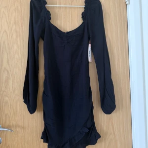 Svart volangklänning från Nelly - Fin svart klänning från Nelly med långa ärmar, volangdetaljer vid axlar och nederkant samt lätt rynk framtill. Klänningen har en v-ringning och dragkedja i ryggen. Aldrig använd och lappen är kvar. 