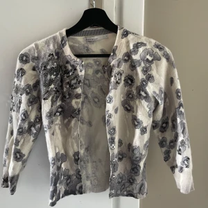 Blommig kofta från Karen Millen - Supersöt kofta från Karen Millen i ljusgrå och vit ton med blommigt mönster och pärlbroderier. Koftan har knappar framtill och är gjord i en mjuk mix av angora, ull och polyamid. Storlek 1 (S).