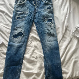 Dsquared2 slitna blå jeans, stl 46 - Riktigt snygga blå jeans från Dsquared2 med distressed look, stora slitningar och hål framtill samt coola färgstänk. Klassisk femficksmodell med knappgylf och Dsquared2-logga bak. Tillverkade i bomull för en skön känsla och streetig vibe. 100% äkta unika 