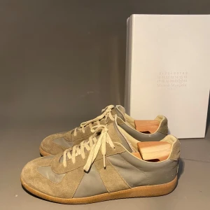 Maison Margiela sneakers i mocka och skinn - Snygga sneakers från Maison Margiela i beige och grå toner. Skorna har en klassisk siluett med ovandel i mocka och skinn, samt beige snörning och gummisula. Perfekta för dig som gillar stilrena och tidlösa sneakers med en lyxig touch.