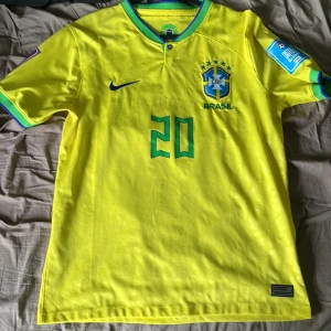 Brasilien VM 2022 Vinicius Jr tröja - Säljer en Brasilien fotbollströja från VM 2022 med Vinicius Jr och nummer 20. Tröjan är gul med gröna och blå detaljer, broderat CBF-märke och Nike-logga. Korta ärmar med gröna och blå muddar, tillverkad i lätt och ventilerande polyester. Tröjan är använd 3-4 ggr! Modellen är 187 cm lång och väger 77 kg.