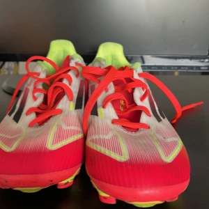 Adidas fotbollsskor i rött och neon f50 - Snygga Adidas fotbollsskor med röd och vit ovandel, neonfärgade detaljer och svarta ränder på sidorna. Skorna har röda snören, gulgrön insida och sula med dobbar för bra grepp på planen. Perfekta för dig som vill sticka ut på matchen!