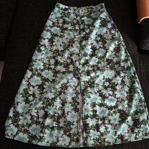 Blommig långkjol från ONLY, strl 36 - Supersnygg långkjol från ONLY med blommigt mönster i ljusblått, grönt och svart. Kjolen har knappar framtill och en lätt utställd passform. Perfekt för dig som vill sticka ut med färg och mönster. Materialet känns lätt och flowy. Den har tappat en knapp men det syns inte så tydligt. 