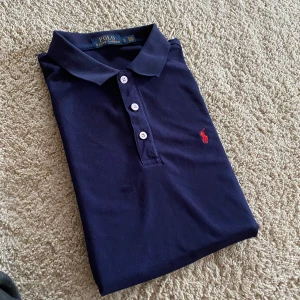 Marinblå pikétröja från Ralph Lauren - Klassisk marinblå pikétröja från Ralph Lauren med röd broderad logga på bröstet. Tröjan har korta ärmar, krage och tre knappar. Perfekt för sommarn.