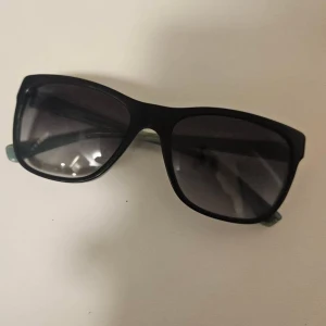 Svarta solglasögon Emporio Armani - Snygga solglasögon från Emporio Armani med svart front och transparent blågrön båge. Rektangulär form och mörka glas ger en cool vibe. Perfekta för att lyfta din stil i sommar. Märkeslogga på insidan av skalmen.